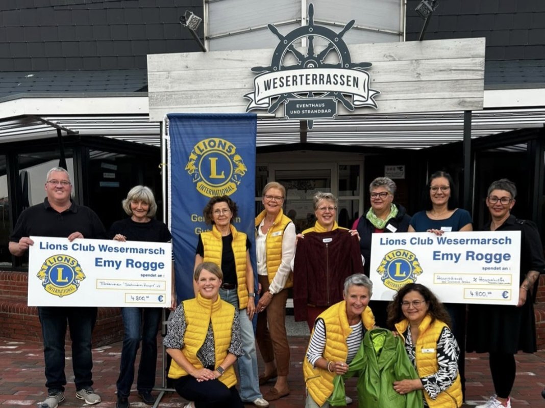 Gruppenfoto vor den Weserterrassen: Mehrere Frauen und ein Mann stehen und knien vor dem Eingang. Einige Frauen tragen gelbe Lions-Westen und halten Kleidungsstücke in den Händen. Zwei Personen halten Spendenschecks des Lions Clubs „Emy Rogge“ in die Kamera.